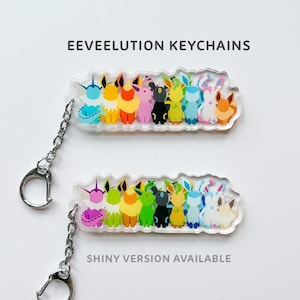 May include: A set of nine clear acrylic keychains featuring the Eevee evolutions. The keychains are arranged in a row, with each evolution depicted in its signature color. The text "EEVEELUTION KEYCHAINS" is printed above the keychains, and the text "SHINY VERSION AVAILABLE" is printed below the keychains.