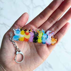Clear acrylic Eeveelution keychain on top of a hand. This small acrylic charm features all the Eeveelutions: Flareon, Jolteon, Vaporeon, Umbreon, Espeon, Leafeon, Glaceon, and Sylveon with a metal clip on attachment.