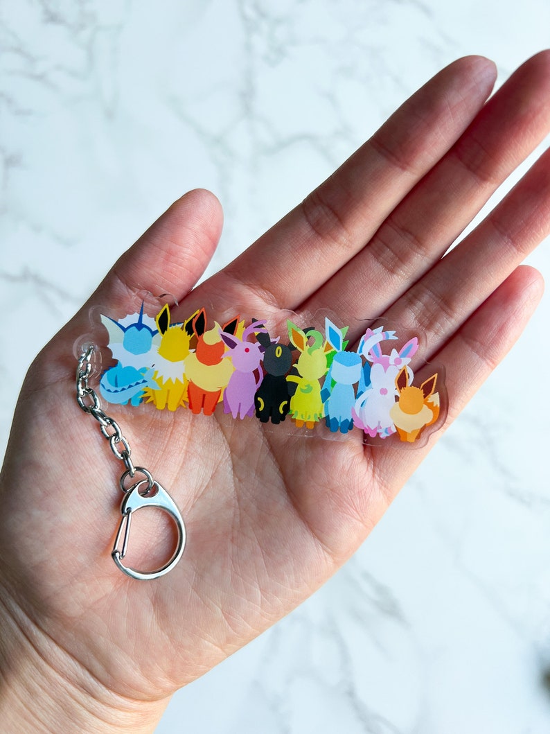 Clear acrylic Eeveelution keychain on top of a hand. This small acrylic charm features all the Eeveelutions: Flareon, Jolteon, Vaporeon, Umbreon, Espeon, Leafeon, Glaceon, and Sylveon with a metal clip on attachment.