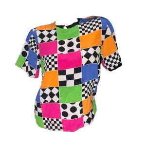 Może przedstawiać: Koszulka z krótkim rękawem z patchworkowym wzorem, w którym znajdują się czarne i białe kwadraty w kratkę, romby, kropki i jednolite kolory w odcieniach różowego, pomarańczowego, zielonego, niebieskiego i białego.