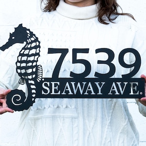 May include: Black metal address sign featuring a seahorse silhouette on the left side. The sign displays the numbers "7539" above the street name "SEA WAY AVE." in a clean, modern font. The sign is held up by a person.