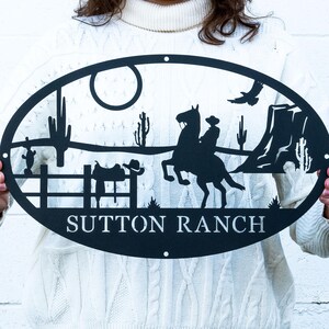 May include: Black metal oval sign featuring a cowboy on horseback, cacti, and the text "SUTTON RANCH." The silhouette design includes a fence, a soaring bird, and a sun. The sign is held up by a person.