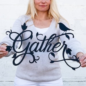 May include: A woman holds a black metal sign with the word "Gather" in a decorative script font. The sign features a bird perched on a branch with leaves.