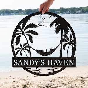 May include: Black metal sign with a silhouette of palm trees, a hammock with a person relaxing in it, and the text "SANDY'S HAVEN".