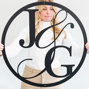 May include: A black metal monogram with the letters "J & G" in a circular design. The letters are stylized and connected, creating a modern and elegant look. The monogram is held up by a person.