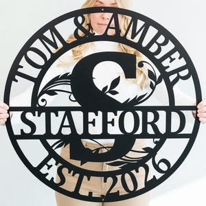 May include: A black metal family name sign featuring the names "TOM & AMBER" and the surname "STAFFORD" in a circular design. The sign also includes the year "EST 2026" and decorative floral elements.