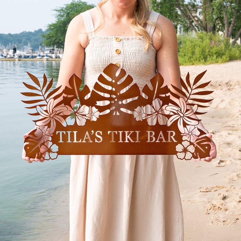 Puede incluir: Un letrero de bar tiki de metal marr&oacute;n con un tema tropical. El letrero presenta hojas de palma recortadas, flores y el texto "TILA'S TIKI BAR".