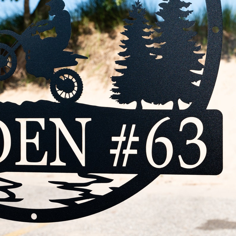Puede incluir: Placa de n&uacute;mero de casa de metal negro con una silueta de una moto de cross y pinos. La placa dice "DEN #63".