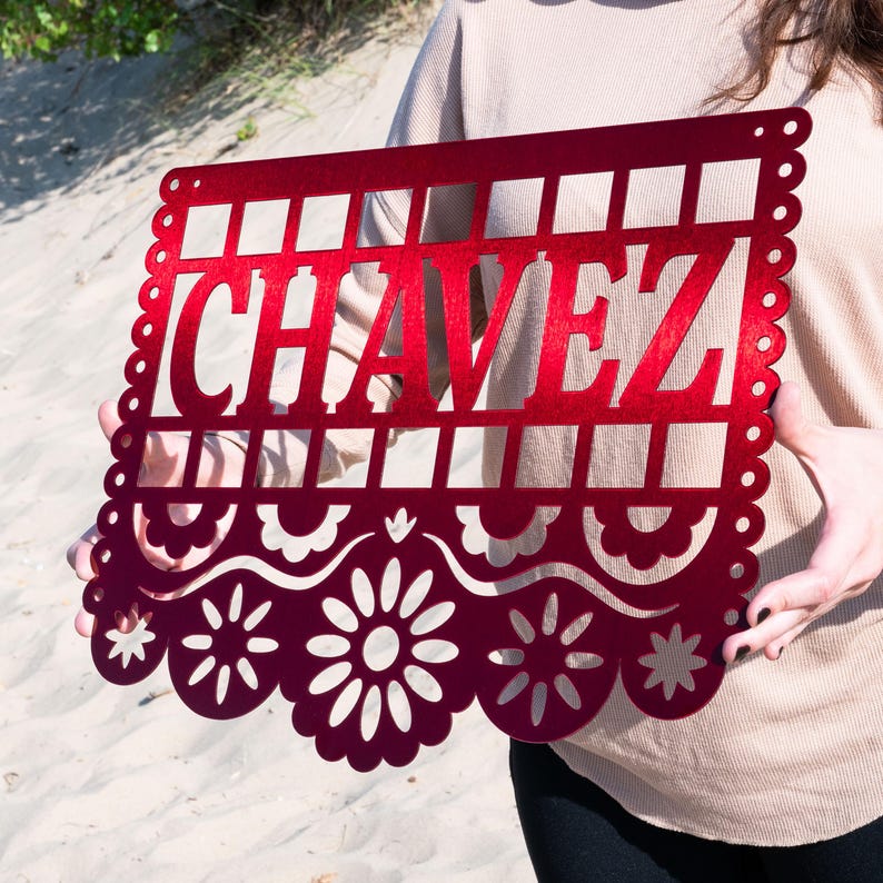 May include: A red metal sign featuring the name "CHAVEZ" in a cut-out design. The sign has a decorative floral and scalloped edge, with a grid pattern above the name. The sign is held up against a sandy background.