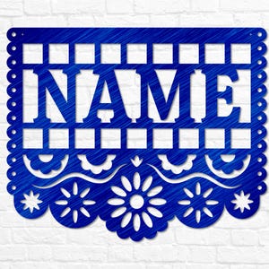 May include: A blue metal sign featuring the word "NAME" in a cut-out design. The sign has a decorative scalloped edge and floral and star cutouts. The background is a white brick wall.