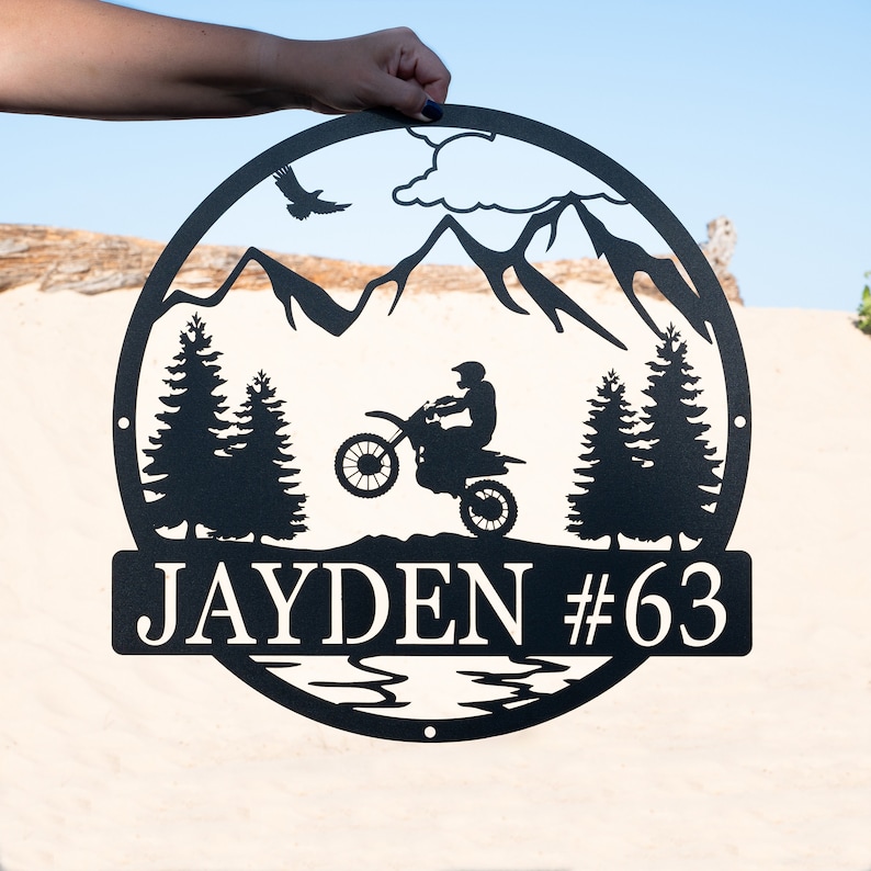 Puede incluir: Se&ntilde;al de metal negro con una escena de monta&ntilde;a, un piloto de motocross y &aacute;rboles. La se&ntilde;al dice "JAYDEN #63".