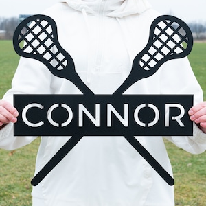 May include: Black metal lacrosse stick silhouette with the name "CONNOR" in white lettering on a black rectangular sign.