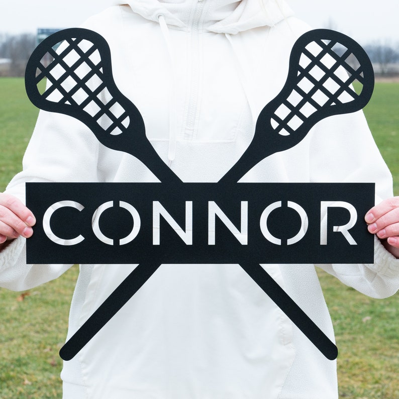 May include: Black metal lacrosse stick silhouette with the name "CONNOR" in white lettering on a black rectangular sign.