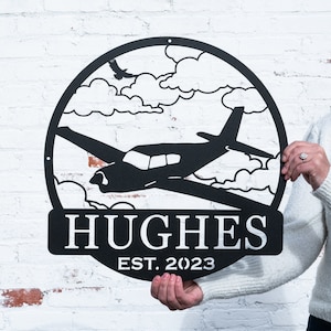 May include: Black metal sign with a silhouette of a small airplane flying through clouds. The sign also includes the text "HUGHES EST. 2023".