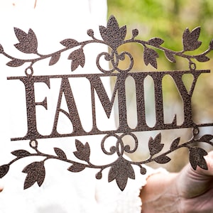 Puede incluir: Un letrero de metal con la palabra "FAMILY" en letras grandes y estilizadas. El letrero est&aacute; decorado con un dise&ntilde;o de vid con hojas y tiene un acabado marr&oacute;n r&uacute;stico.