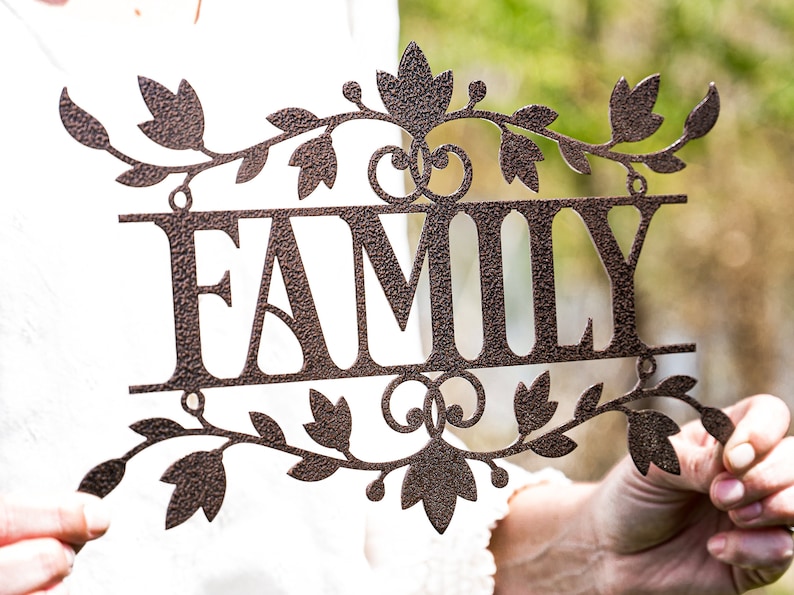 Puede incluir: Un letrero de metal con la palabra "FAMILY" en letras grandes y estilizadas. El letrero est&aacute; decorado con un dise&ntilde;o de vid con hojas y tiene un acabado marr&oacute;n r&uacute;stico.