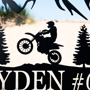 Puede incluir: Silueta negra de metal de una persona montando una moto de cross, con dos pinos a cada lado de la moto. El nombre "RYDEN" y el n&uacute;mero "#" son visibles en la parte inferior de la imagen.