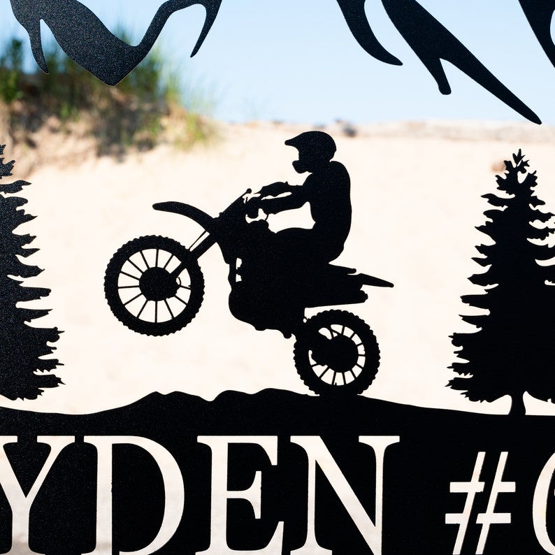 Puede incluir: Silueta negra de metal de una persona montando una moto de cross, con dos pinos a cada lado de la moto. El nombre "RYDEN" y el n&uacute;mero "#" son visibles en la parte inferior de la imagen.