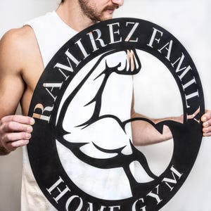 K&ouml;nnte beinhalten: Schwarzes Metallschild mit einer wei&szlig;en Silhouette eines Bizepses und dem Text "RAMIREZ FAMILY HOME GYM" in einem Kreis.