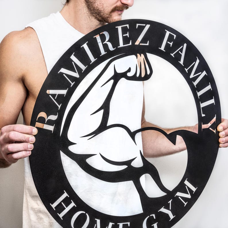 K&ouml;nnte beinhalten: Schwarzes Metallschild mit einer wei&szlig;en Silhouette eines Bizepses und dem Text "RAMIREZ FAMILY HOME GYM" in einem Kreis.