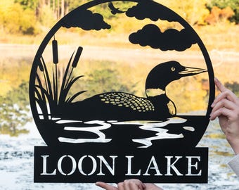 May include: Black metal silhouette of a loon swimming in a lake with reeds and clouds. The text "LOON LAKE" is below the image.