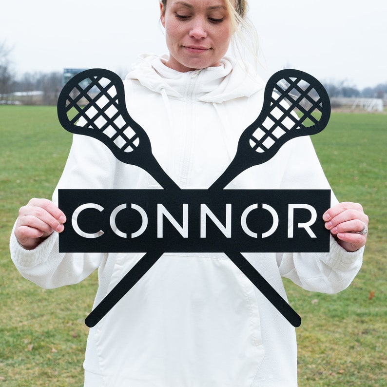 May include: Black metal lacrosse stick sign with the name "CONNOR" in white lettering.