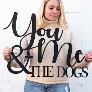 May include: Black metal sign featuring the text "You & Me THE DOGS" in a decorative script. The sign is held by a person wearing a beige sweater and blue jeans, set against a white brick wall.