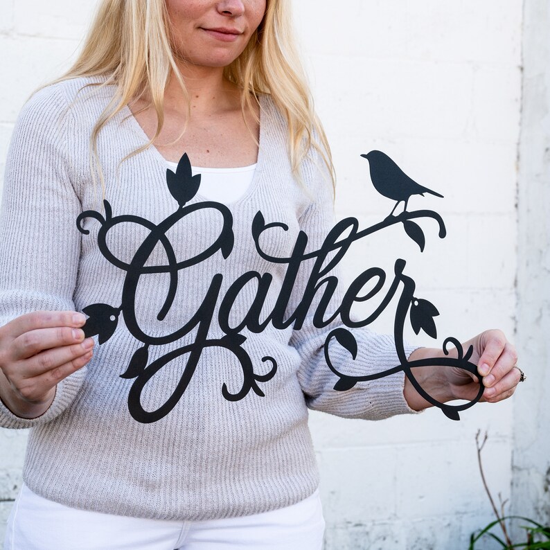 May include: Black metal wall decor featuring the word "Gather" in a decorative script font. The word is surrounded by stylised leaves and a small bird perched on a branch.