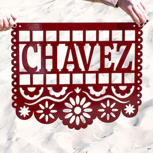May include: A deep red metal sign featuring the name "CHAVEZ" in cut-out capital letters. The sign has a decorative border with a scalloped edge and floral patterns. The sign is held up by two hands.