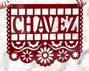 May include: A burgundy metal sign with the name "CHAVEZ" cut out in large capital letters. The sign has a decorative border with scalloped edges and floral designs. The sign is held up by two hands.