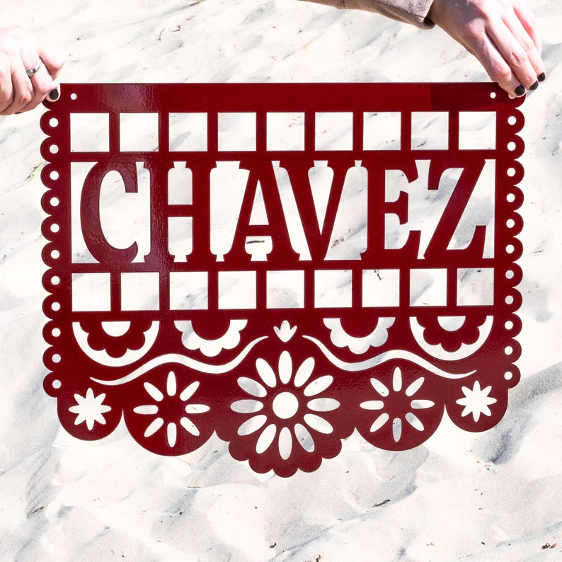 May include: A deep red metal sign featuring the name "CHAVEZ" in cut-out capital letters. The sign has a decorative border with a scalloped edge and floral patterns. The sign is held up by two hands.
