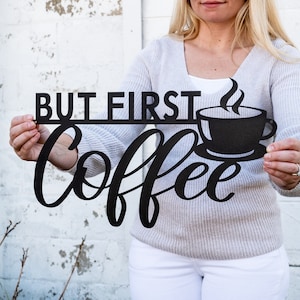 May include: Black metal sign with the text "But First Coffee" in a cursive font. A stylized coffee cup with steam rising from it is above the word "Coffee".