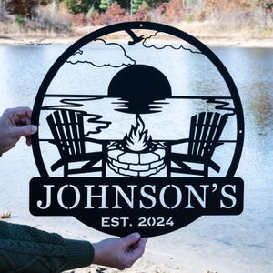 May include: Black metal sign with a silhouette of a lake scene with two chairs, a fire pit, and a setting sun. The sign reads "JOHNSON'S EST. 2024".