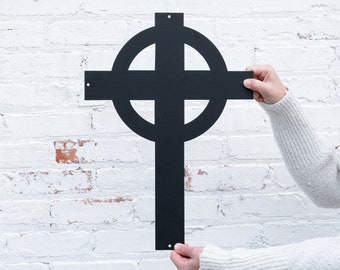 May include: A black metal Celtic cross with a circle design. The cross is being held up by a person's hands.