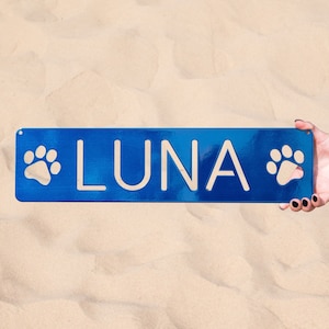 May include: A blue metal sign with paw prints on either side and the name "LUNA" in white lettering.