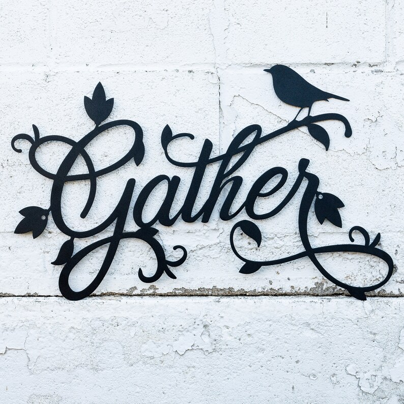 May include: Black metal wall decor featuring the word "Gather" in a decorative script font. The word is surrounded by stylised floral designs and a small bird silhouette.