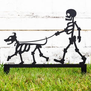 May include: Black metal silhouette of a skeleton walking a dog. The skeleton is wearing a simple skeletal costume and the dog is also a skeleton. The silhouette is mounted on a black metal rod.