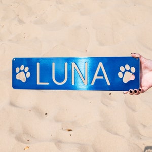 May include: A blue metal sign with the name "LUNA" in white lettering and a paw print on each side.