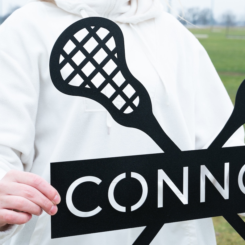 May include: Black metal lacrosse stick silhouette with the word "CONN" cut out in the center of the sign. The sign is held by a person wearing a white hoodie.