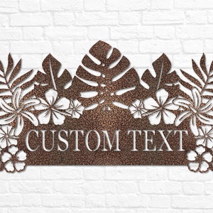 Puede incluir: Un letrero de metal con tem&aacute;tica tropical que presenta una gran hoja de monstera, flores de hibisco y hojas de palma. El letrero tiene un espacio para texto personalizado, que dice "CUSTOM TEXT".