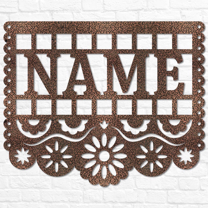 May include: A copper-coloured metal sign with the word "NAME" cut out in capital letters. The sign has a decorative scalloped edge and floral and geometric cut-out patterns. It is displayed against a white brick wall.