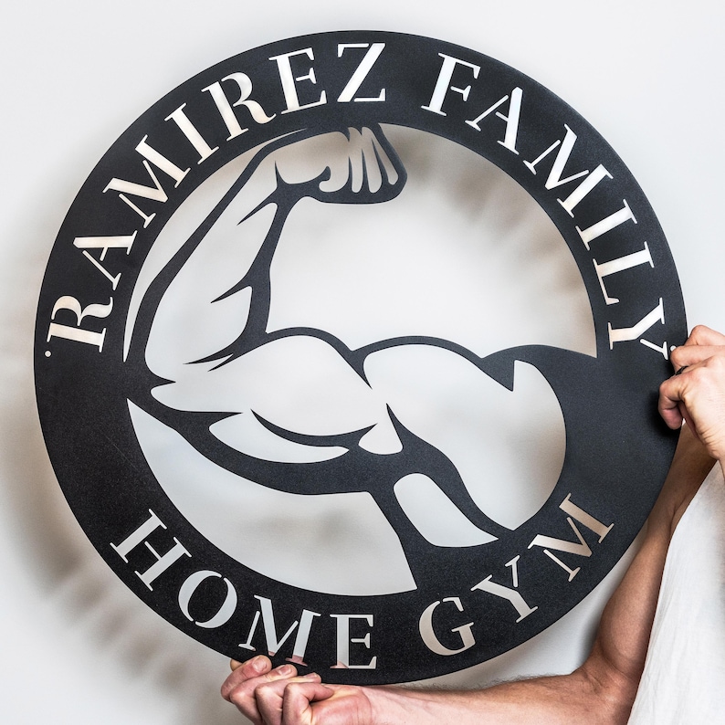 K&ouml;nnte beinhalten: Schwarzes Metallschild mit einer wei&szlig;en Silhouette eines sich anstrengenden Bizepses. Das Schild tr&auml;gt die Aufschrift "Ramirez Family Home Gym".