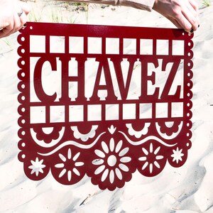May include: A red metal sign featuring the name "CHAVEZ" in a cut-out design. The sign has a decorative scalloped border and floral and geometric patterns. It is held up against a sandy background, showcasing its intricate details.