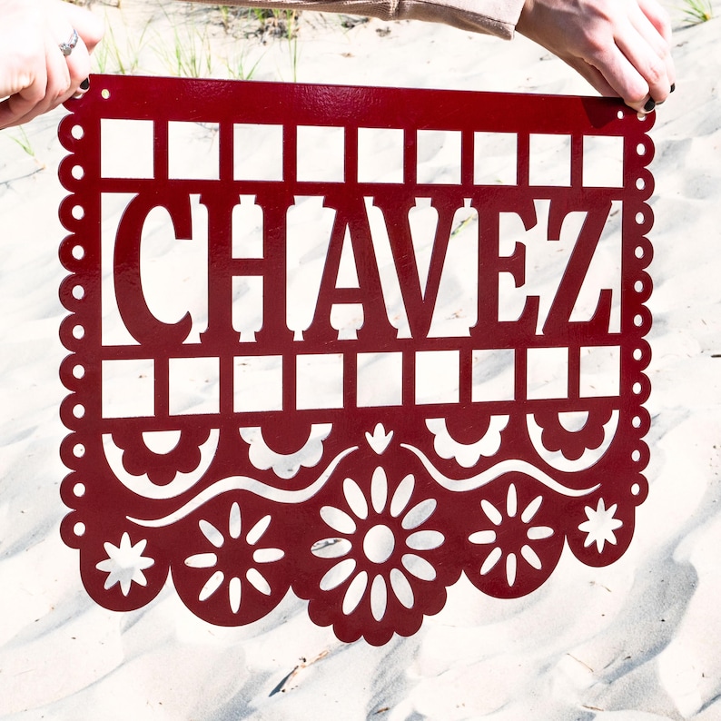 May include: A red metal sign featuring the name "CHAVEZ" in a cut-out design. The sign has a decorative scalloped border and floral and geometric patterns. It is held up against a sandy background, showcasing its intricate details.