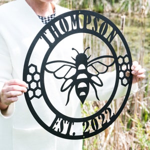 May include: Black metal sign with a bee silhouette in the center. The sign reads "Thompson Est. 2022" around the bee.