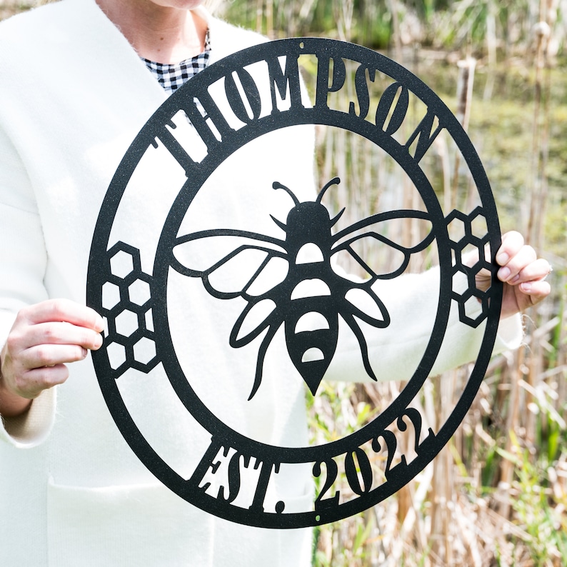 May include: Black metal sign with a bee silhouette in the center. The sign reads "Thompson Est. 2022" around the bee.