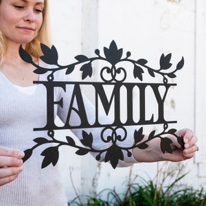 Puede incluir: Un letrero de metal negro con la palabra "FAMILY" en letras grandes rodeado de dise&ntilde;os florales decorativos.