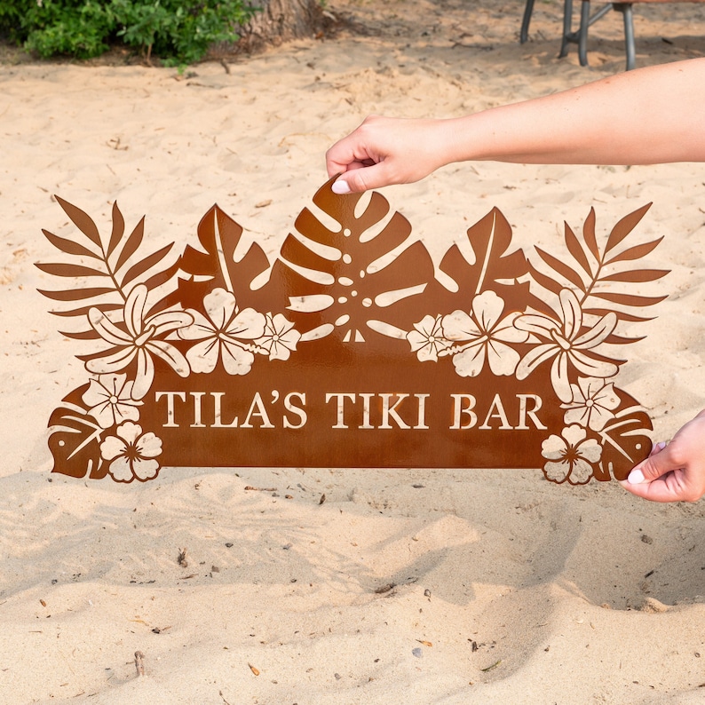 Puede incluir: Un letrero de metal marr&oacute;n oxidado con un tema tropical. El letrero presenta un borde de hojas con flores y el texto "TILA'S TIKI BAR".