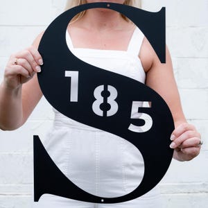 May include: Black metal letter S with the number 185 cut out in white. The letter is being held by a person.