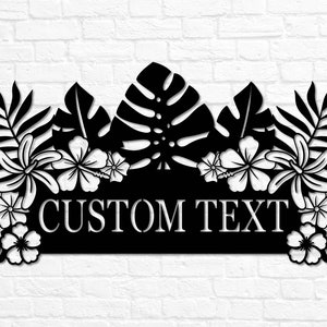 Puede incluir: Letrero de metal negro con tem&aacute;tica tropical con hojas de palma, flores de hibisco y el texto "CUSTOM TEXT".